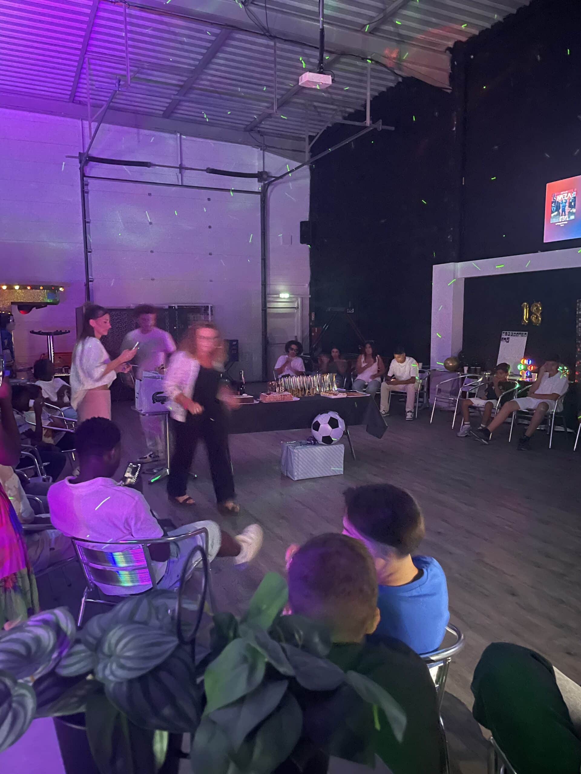 Fête de 18e anniversaire avec gâteau, ballon de football et invités sous des lumières disco violettes.