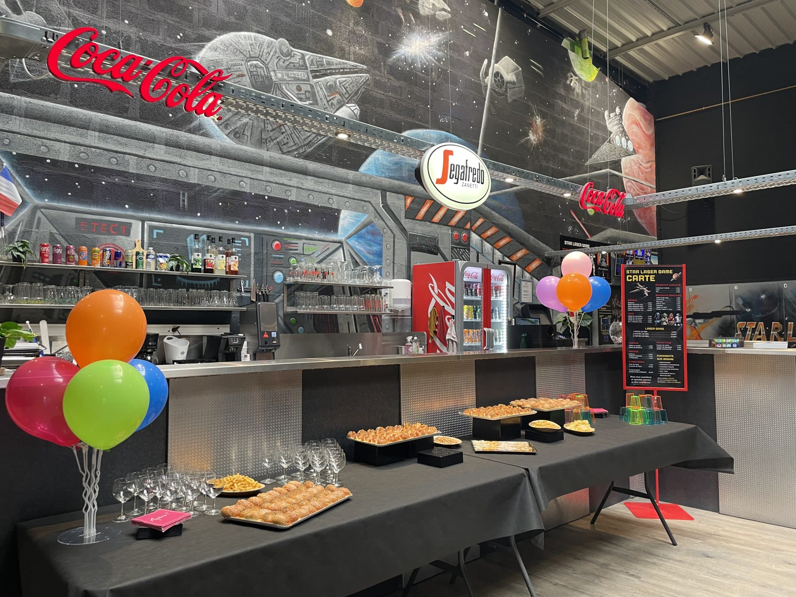 Buffet d'amuse-bouches au bar du Star Laser Game avec ballons colorés et fresque Star Wars murale Coca-Cola.