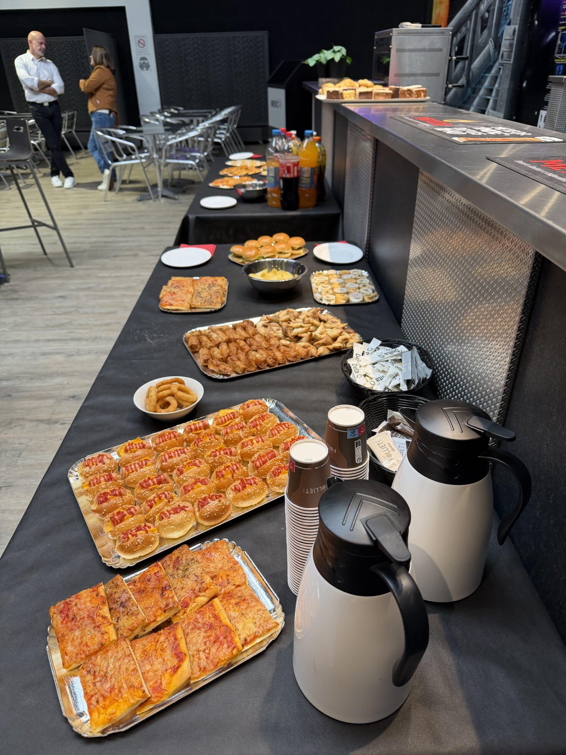 Buffet varié avec mini hot-dogs, pizzas, samoussas et carafes de café pour un événement professionnel convivial.