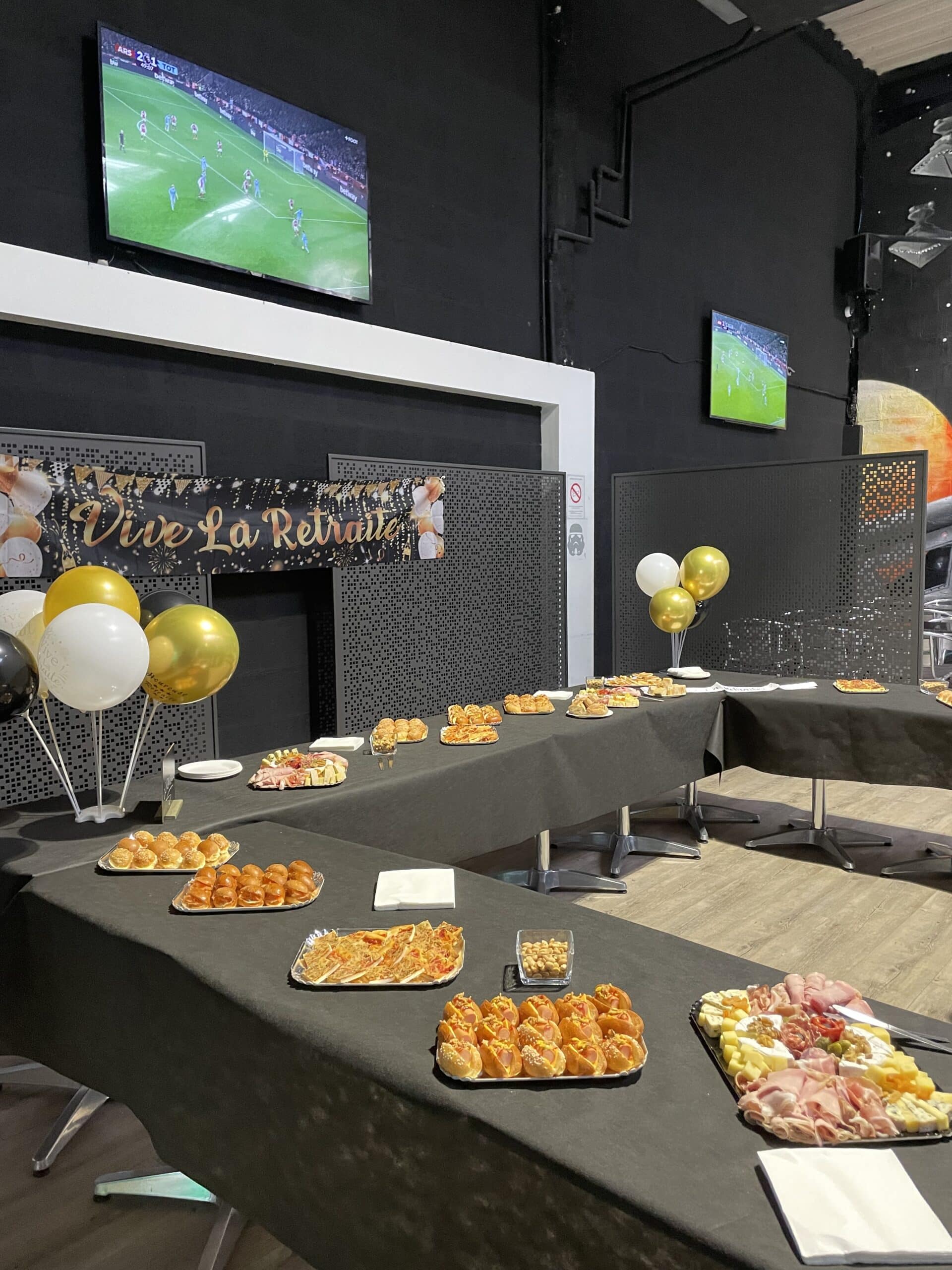 Buffet festif Vive La Retraite avec mini burgers, charcuterie et ballons devant un match de football à la télé.