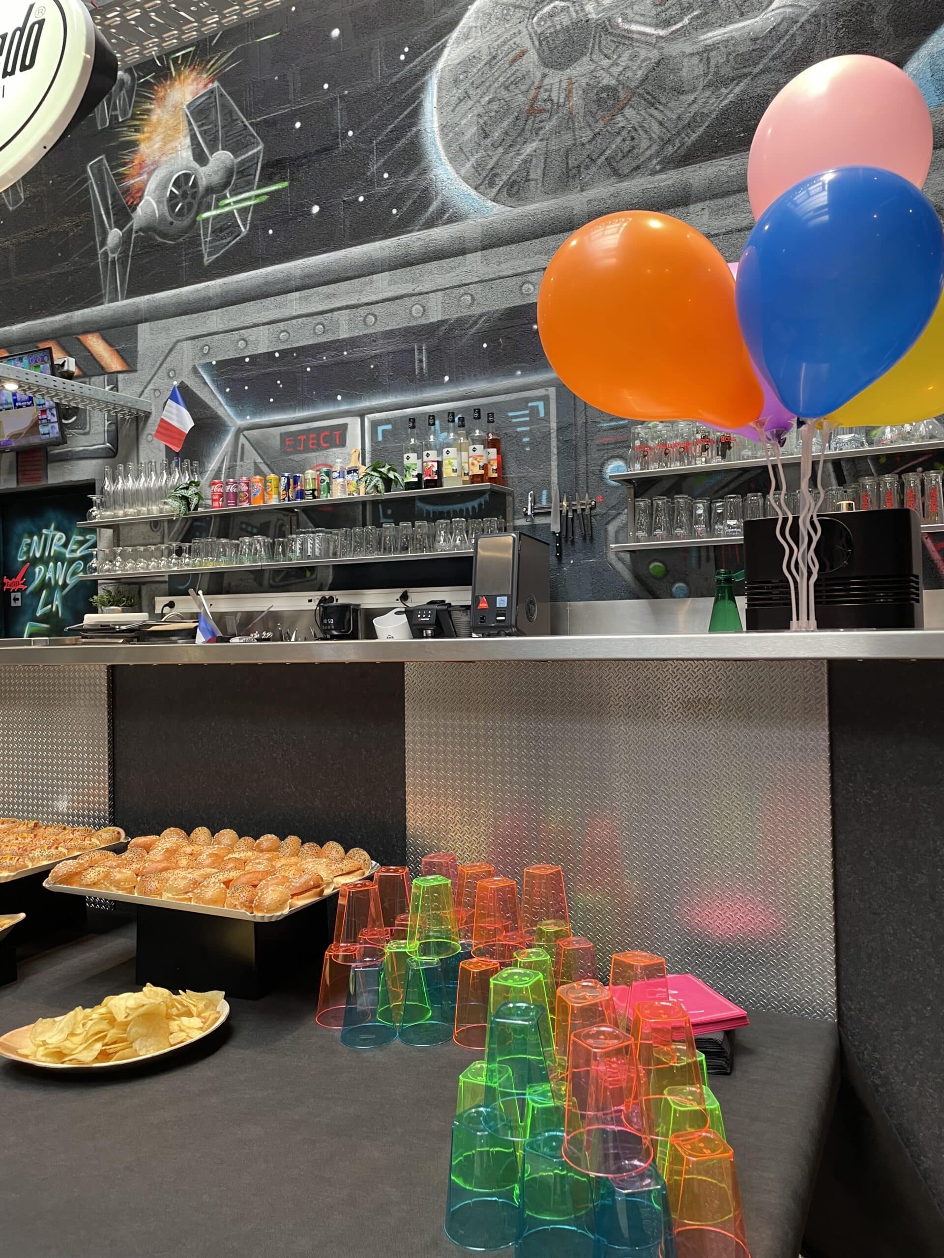 Buffet de fête sur le thème Star Wars avec mini-burgers, chips, verres néon et ballons colorés devant un bar.