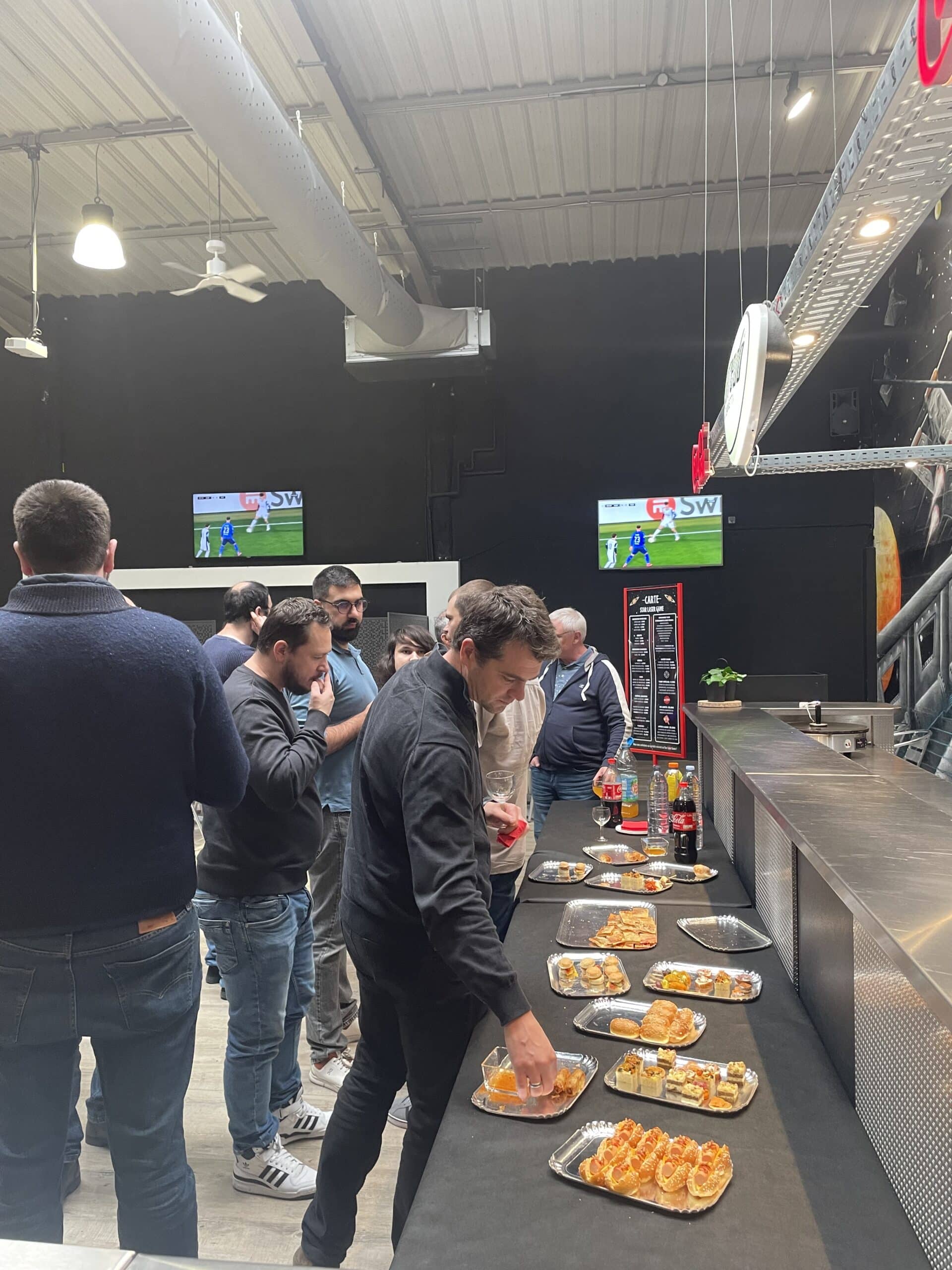 Buffet d'amuse-bouches et menu laser game dans un bar sportif avec écrans diffusant un match de football.
