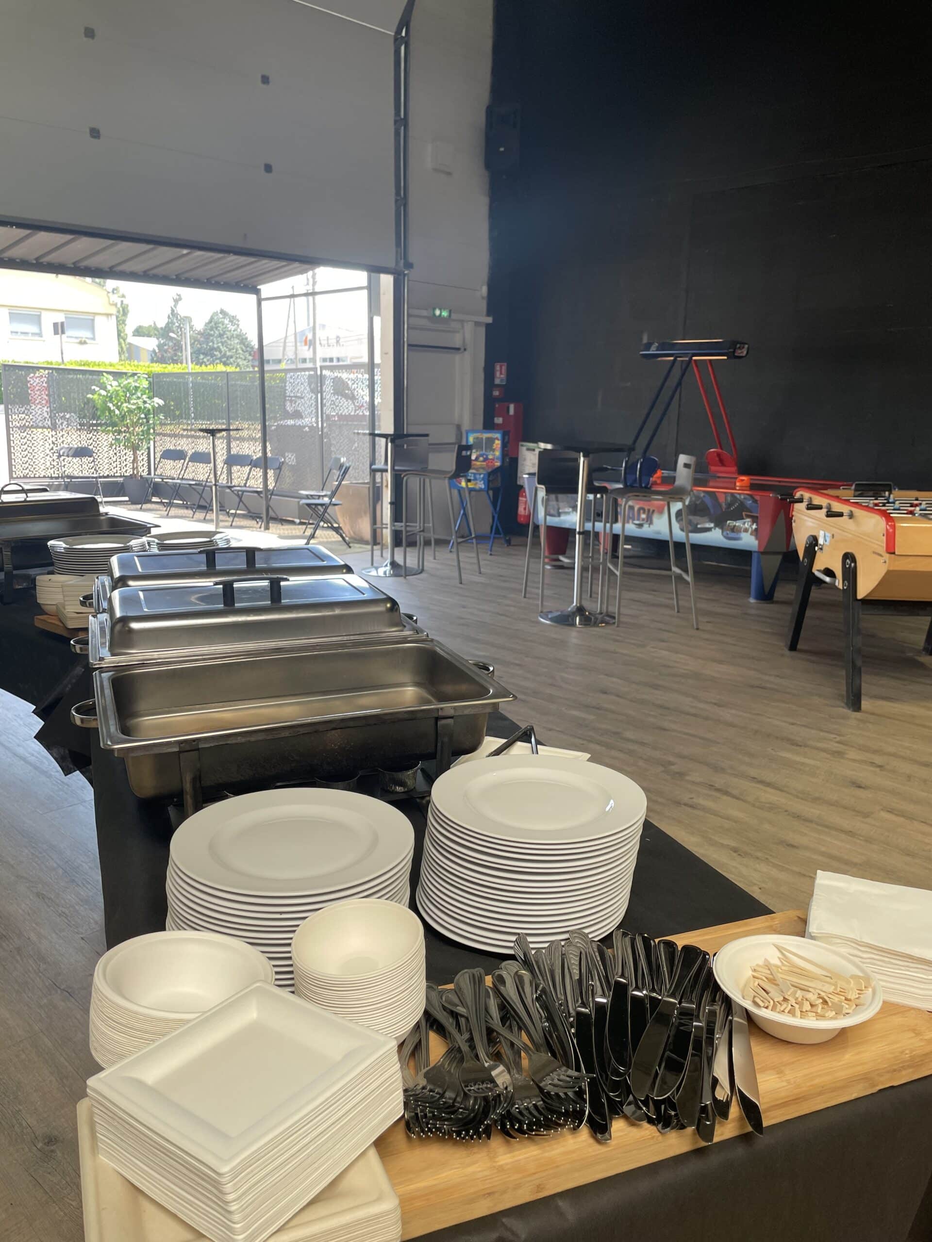 Buffet traiteur avec assiettes et plats chauffants devant une salle de jeux avec baby-foot et air-hockey.