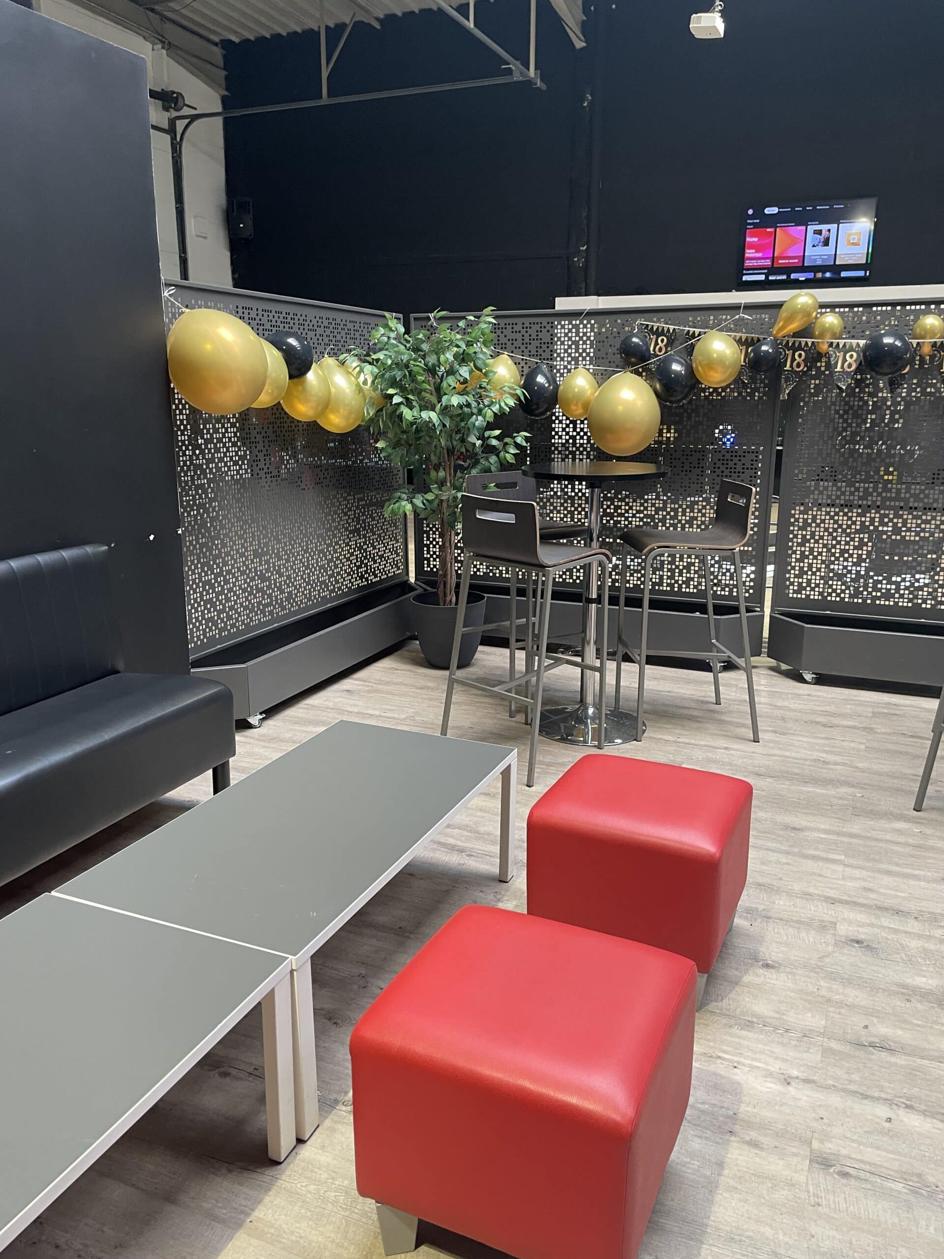 Salon lounge décoré pour un 18ème anniversaire avec ballons noirs et or, banquette noire et poufs rouges.