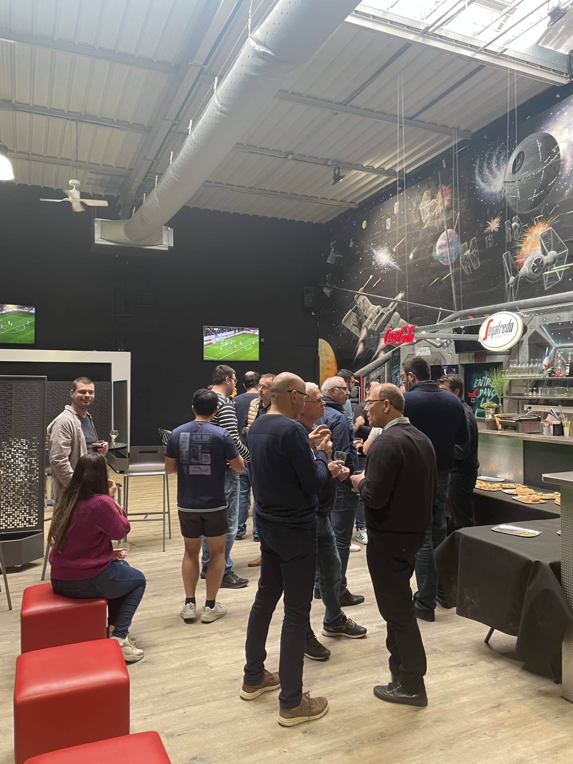 Événement d'entreprise dans une salle avec fresque Star Wars, buffet, et écrans diffusant un match de football.
