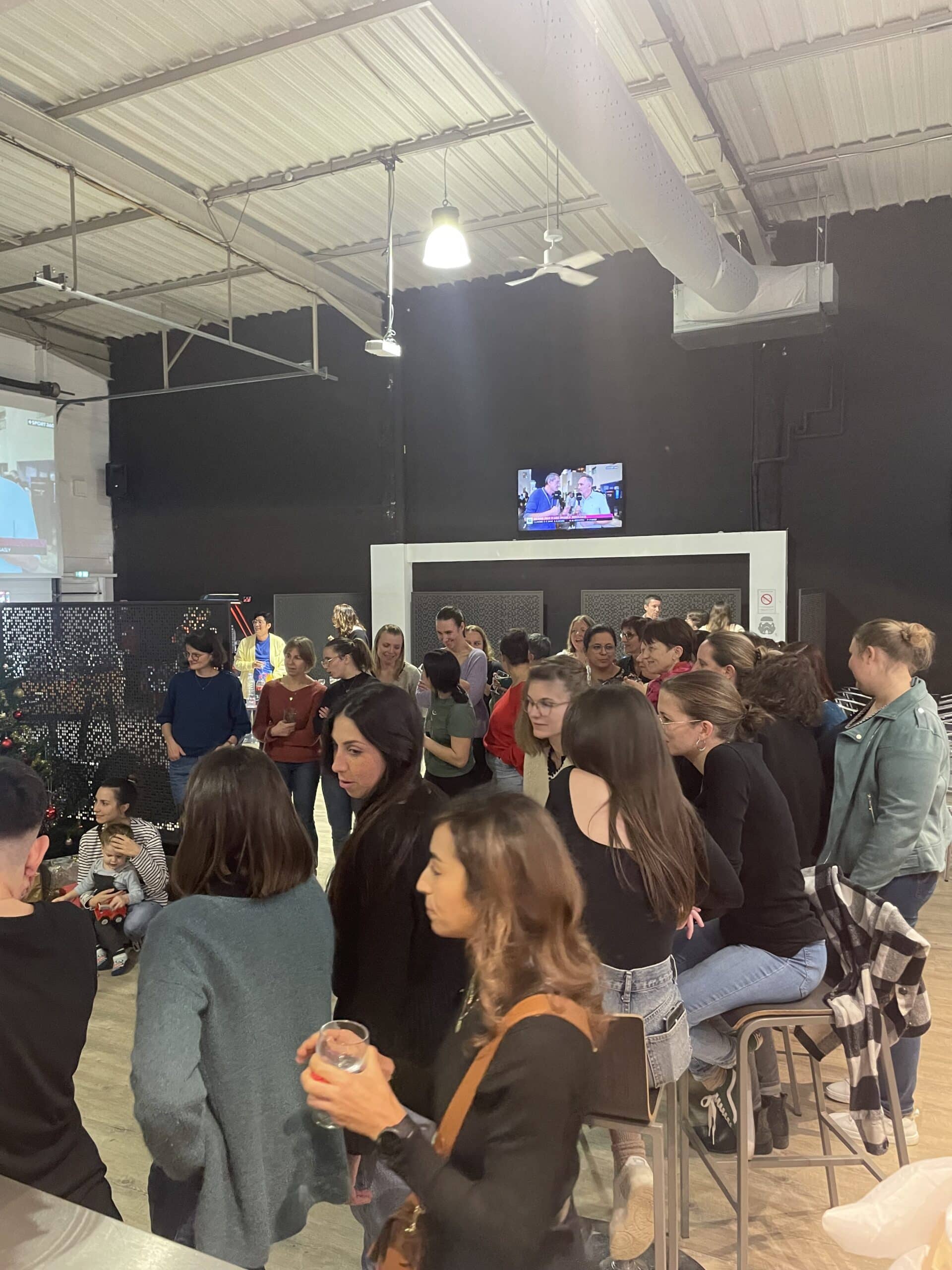Groupe de personnes lors d'un événement social dans une salle industrielle avec écrans de diffusion.
