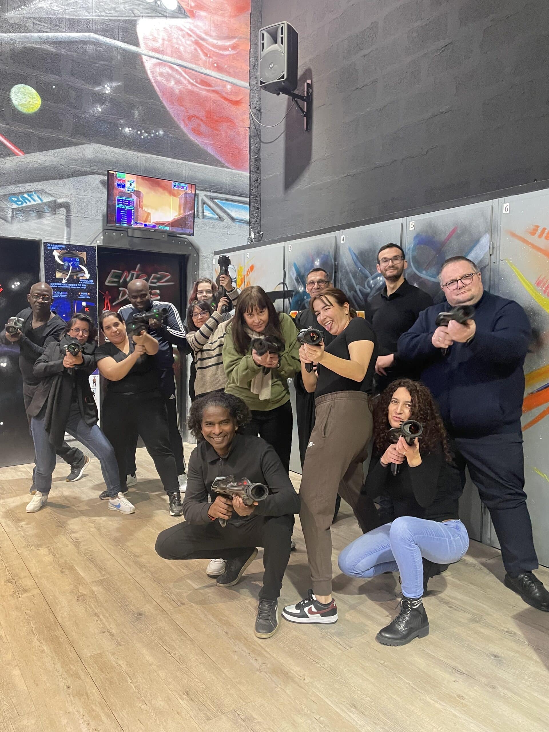 Groupe souriant posant avec des pistolets laser tag dans une salle de jeu spatiale avec graffitis et ambiance dynamique.