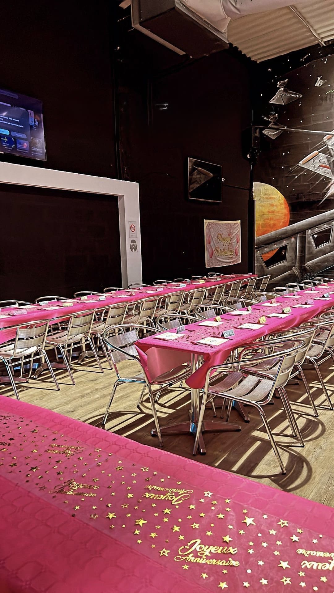 Salle d'anniversaire avec nappes roses Joyeux Anniversaire, chaises en métal et décoration murale thème espace.