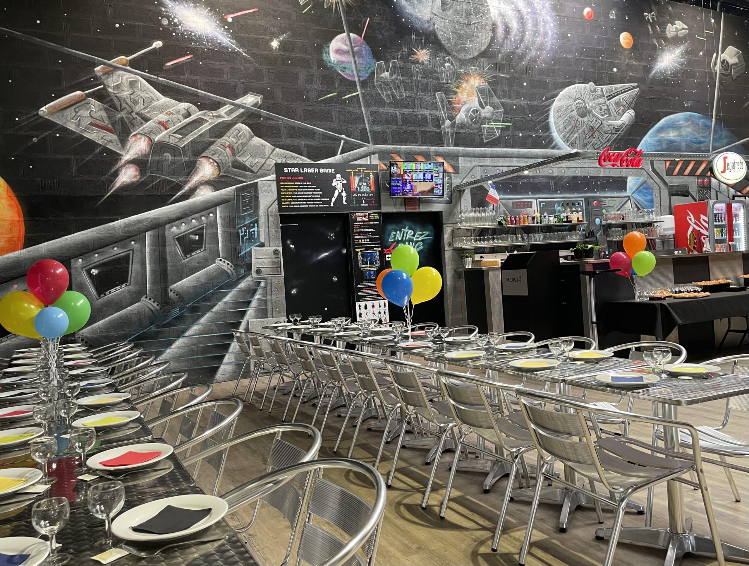 Salle de fête Star Laser Game avec fresque murale Star Wars, ballons colorés et tables dressées pour un repas d'anniversaire.