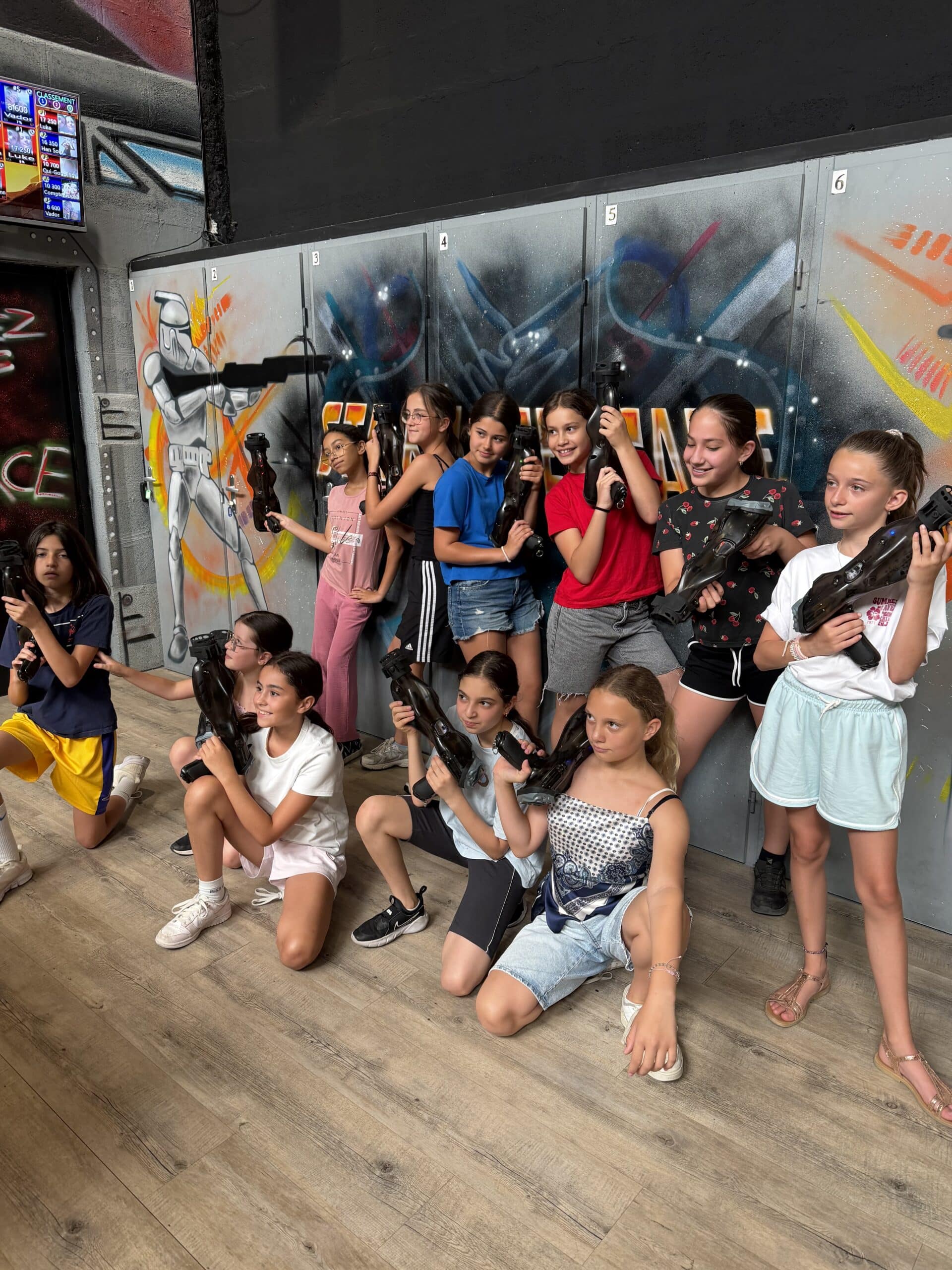 Groupe de filles souriantes avec pistolets laser tag devant un mur graffiti et un écran de classement.
