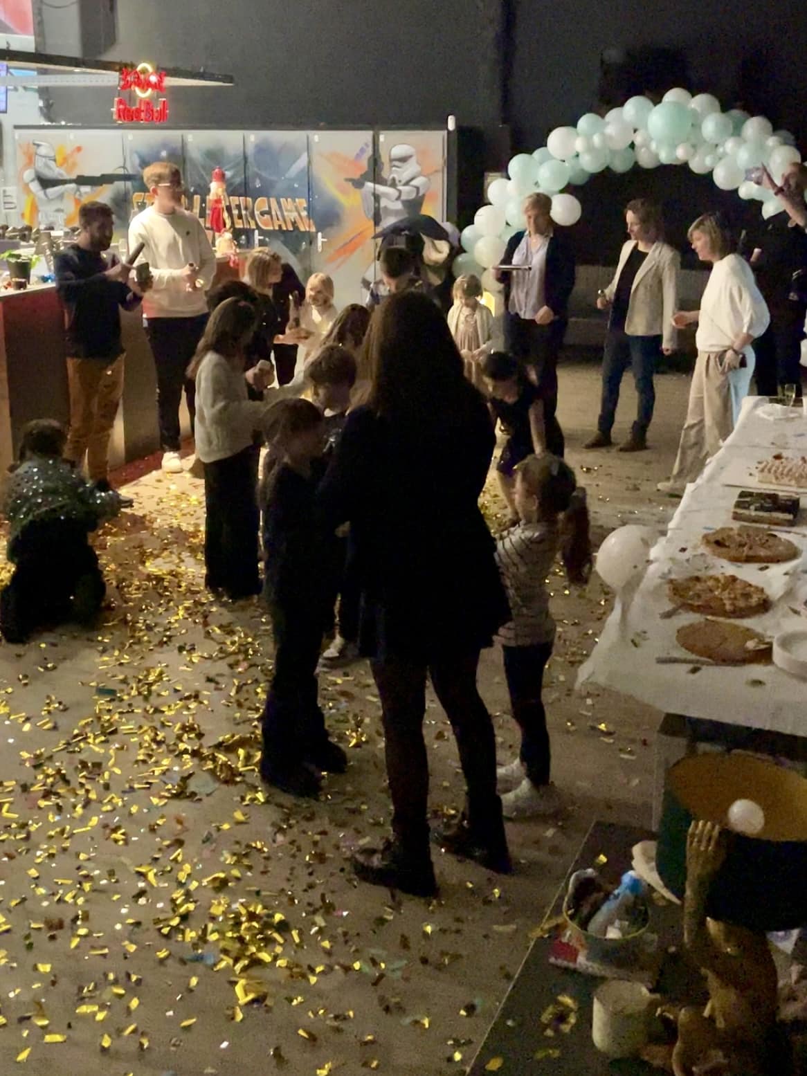 Fête d'anniversaire dans un laser game avec enfants, adultes, confettis dorés au sol et buffet gourmand.