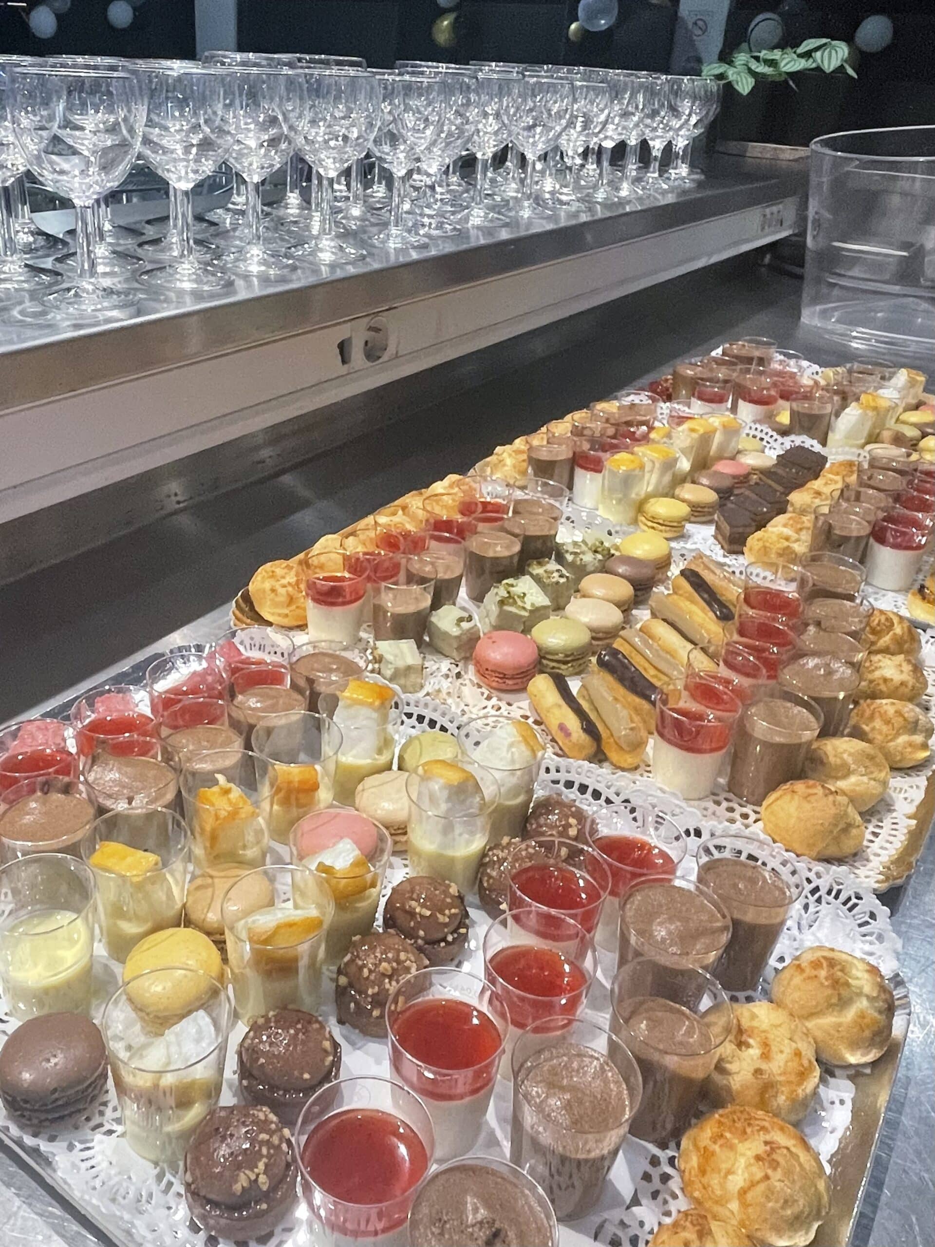 Buffet de mignardises avec verrines colorées, macarons et éclairs devant des rangées de verres à vin élégants.