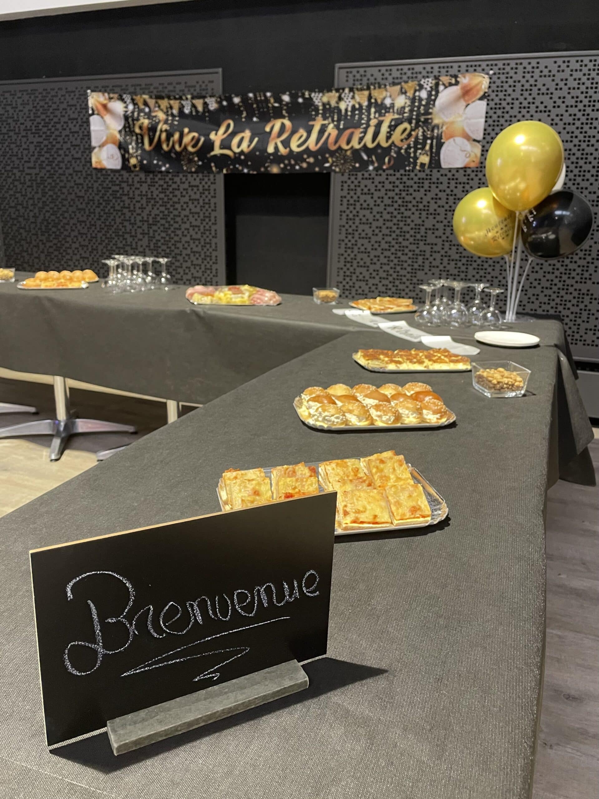 Buffet festif avec panneau Bienvenue, banderole Vive La Retraite, ballons dorés et amuse-bouches sur table.