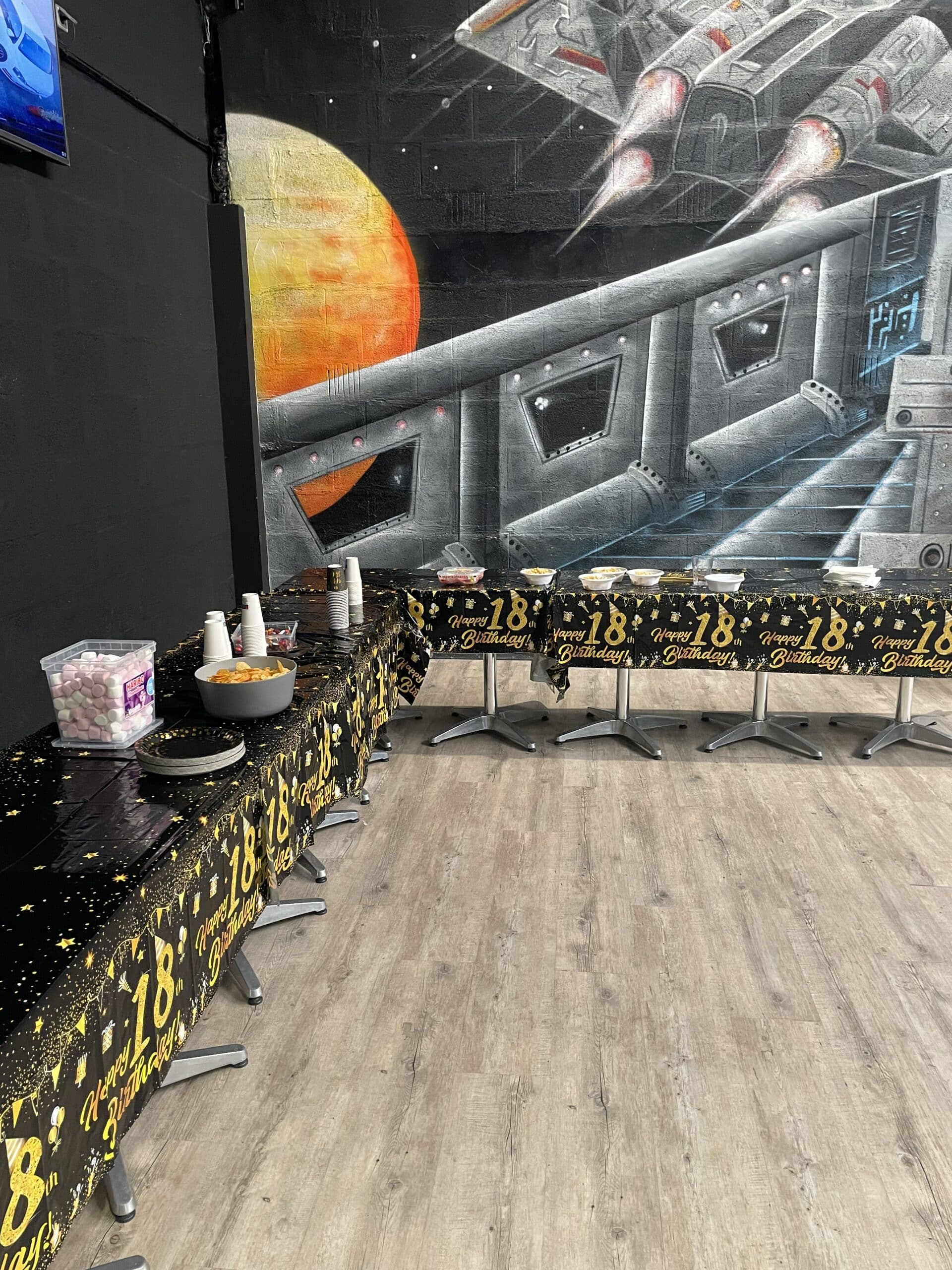 Table de fête pour un 18ème anniversaire avec nappe noire et dorée, buffet et fresque spatiale en arrière-plan.