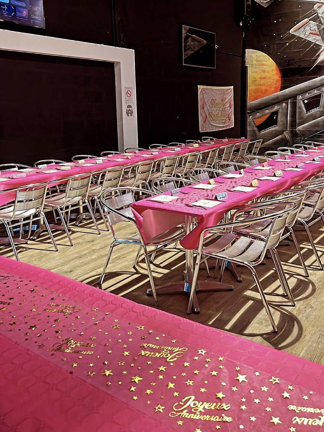 Tables de fête avec nappes roses, chaises argentées et décoration Joyeux Anniversaire dans une salle à thème spatial.