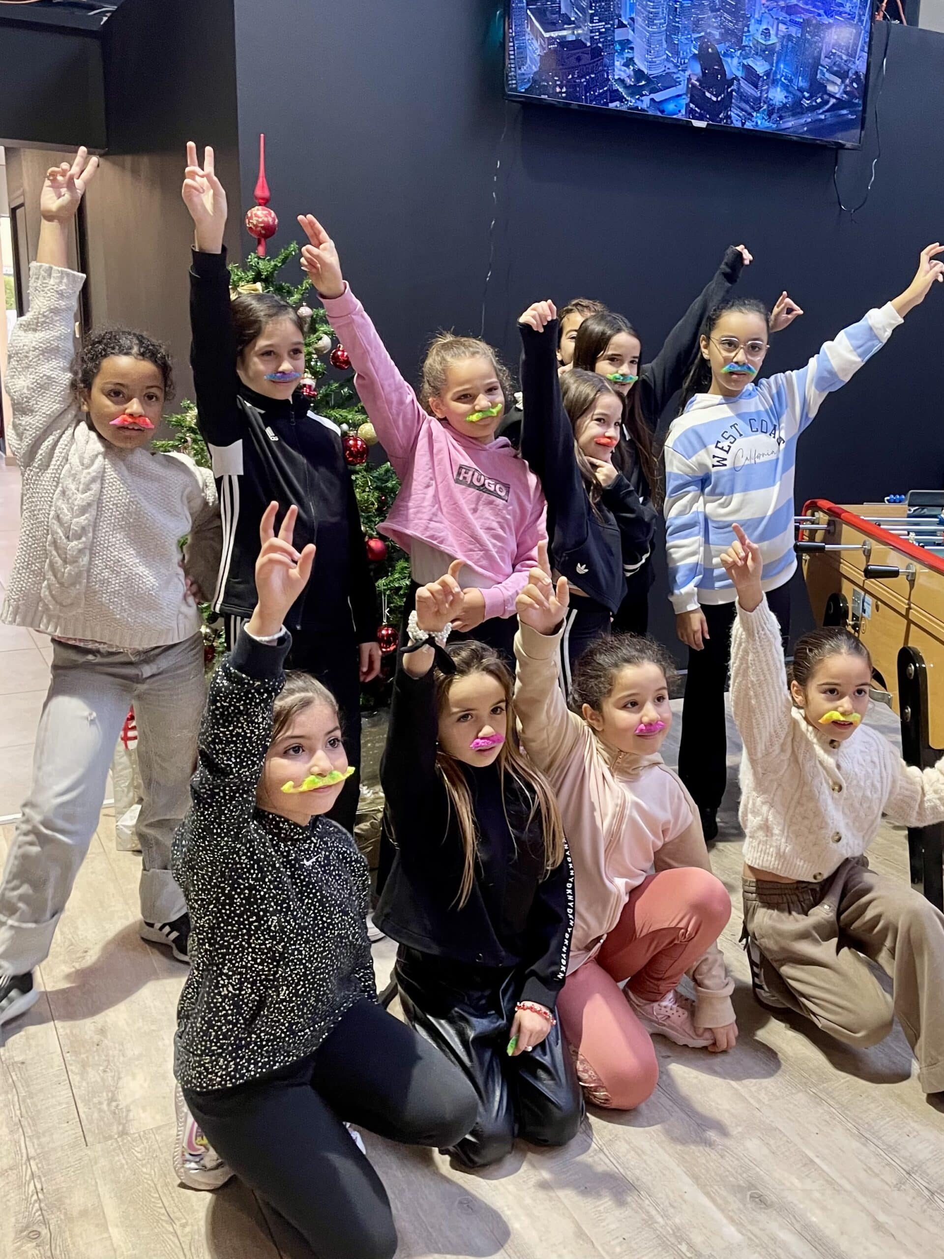 Groupe de jeunes filles avec des moustaches bonbons colorées levant la main devant un sapin de Noël.