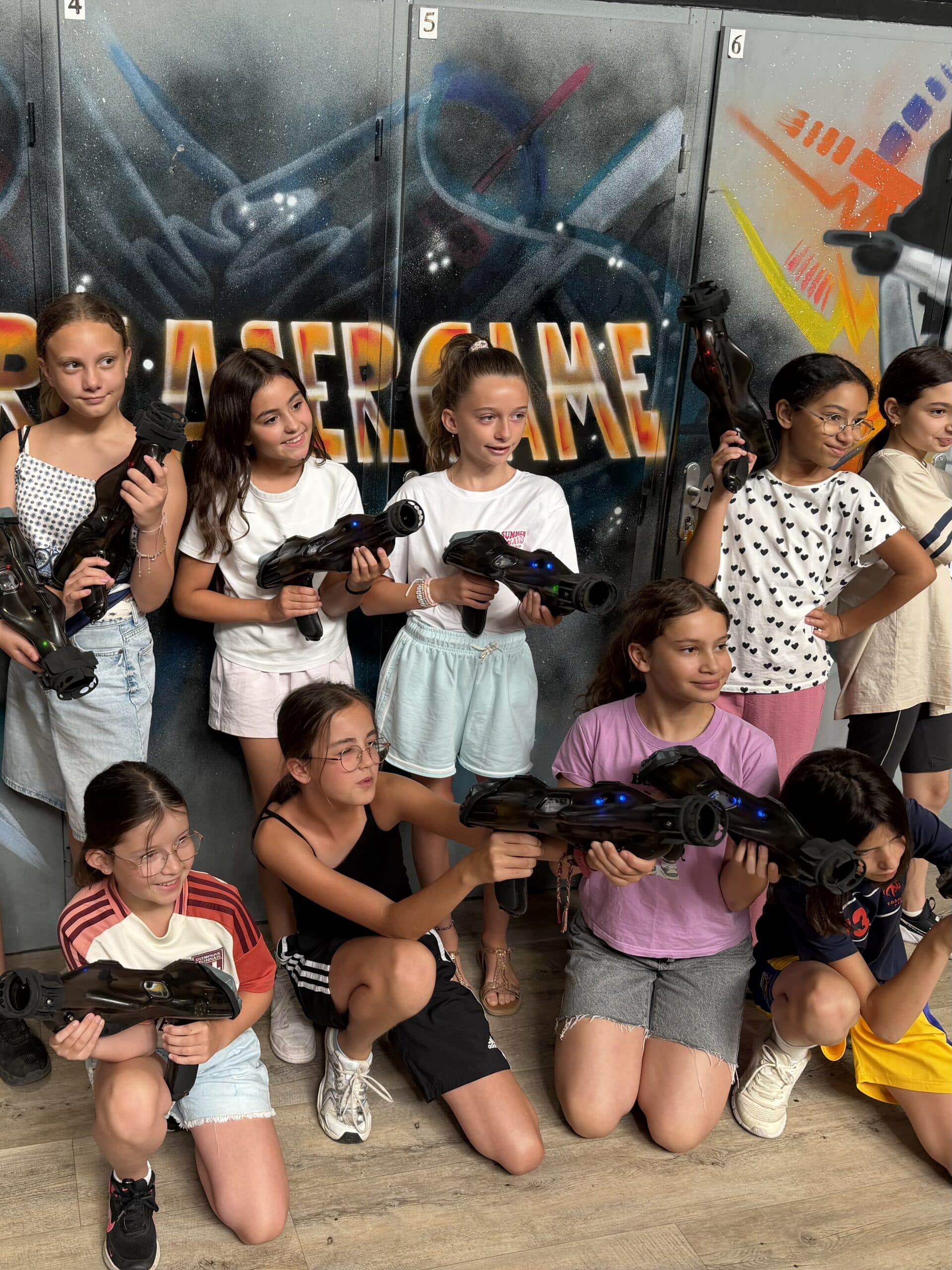 Groupe de jeunes filles souriantes tenant des pistolets de laser game devant un mur tagué LASERGAME.