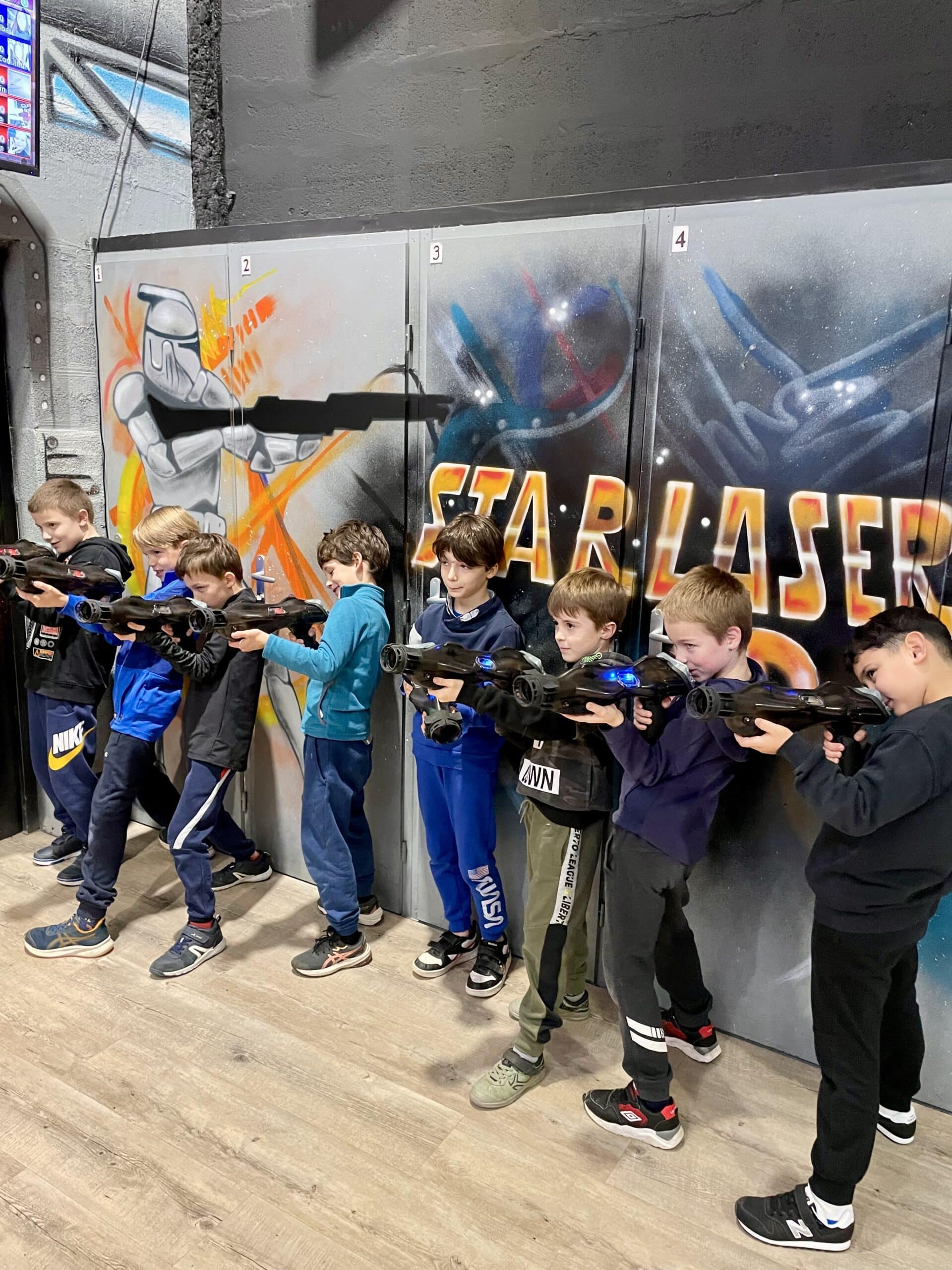 Groupe de garçons jouant au laser tag devant une fresque Star Laser, visant avec leurs équipements avec concentration.