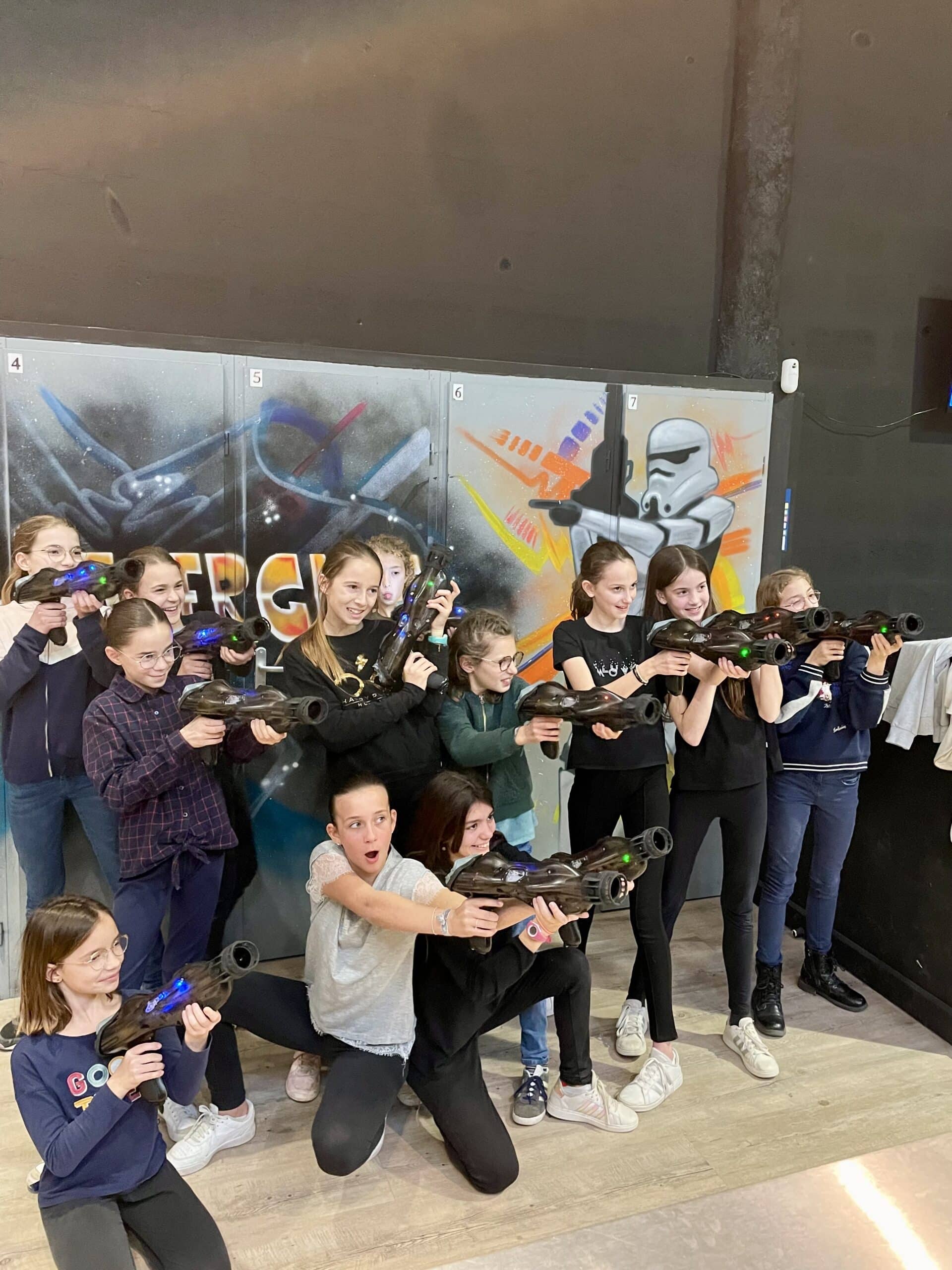 Jeunes filles souriantes visant avec des pistolets laser tag lors d'une session de jeu en groupe devant un mur décoré.
