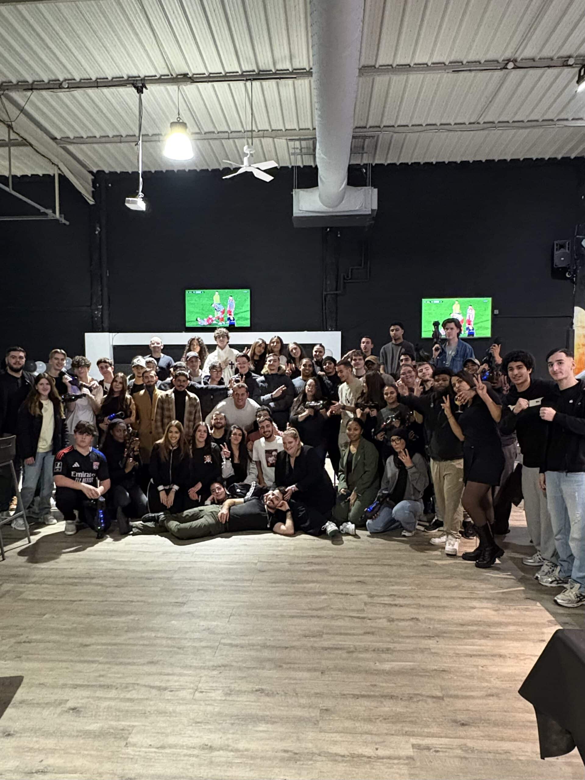 Groupe souriant posant pour une photo lors d'un événement festif dans une salle spacieuse avec écrans de football.