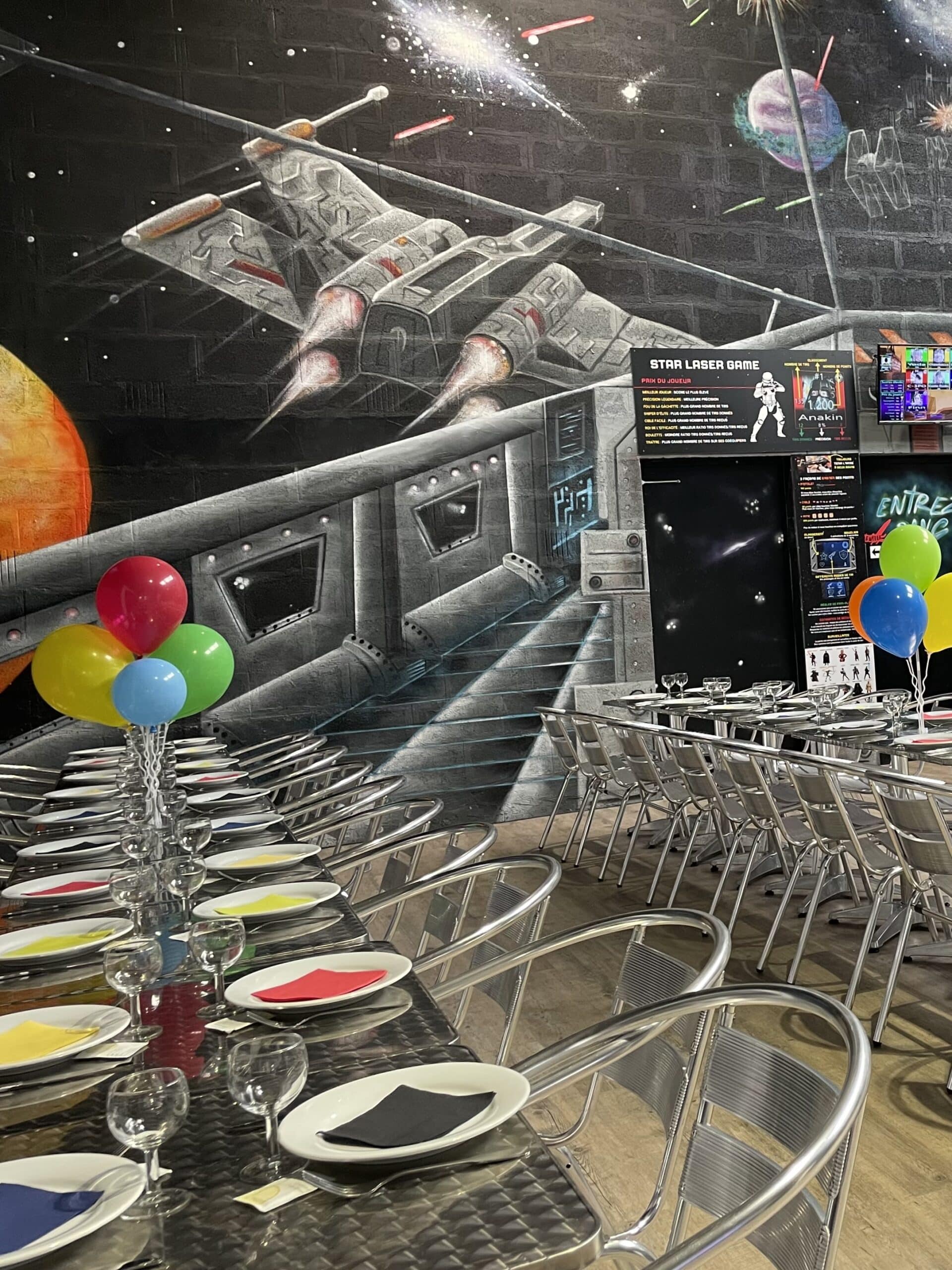 Salle de fête Star Laser Game avec fresque spatiale, ballons colorés et longues tables dressées pour un anniversaire.