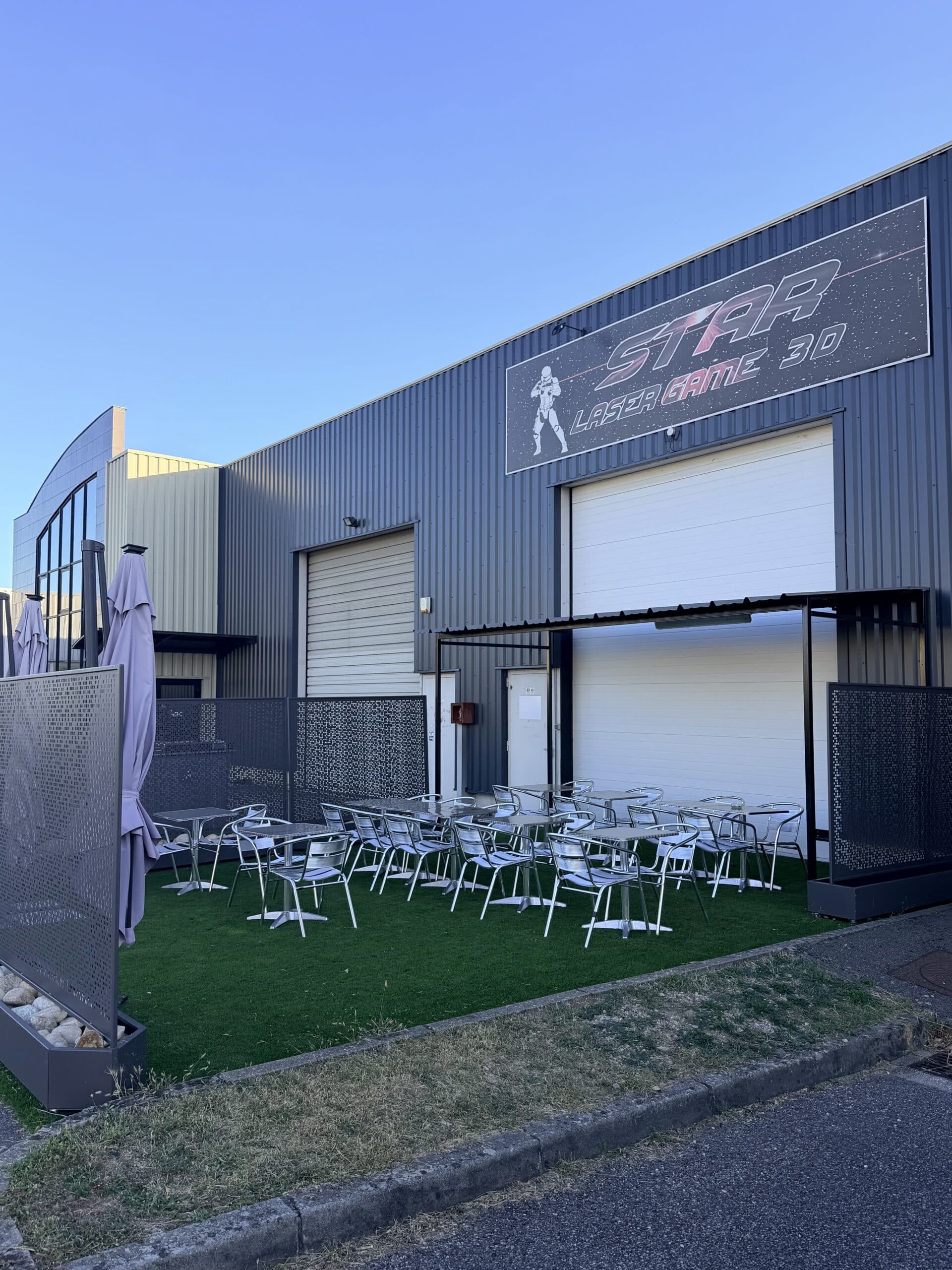Entrée du Star Laser Game 3D avec sa terrasse aménagée de tables en métal sur gazon synthétique.